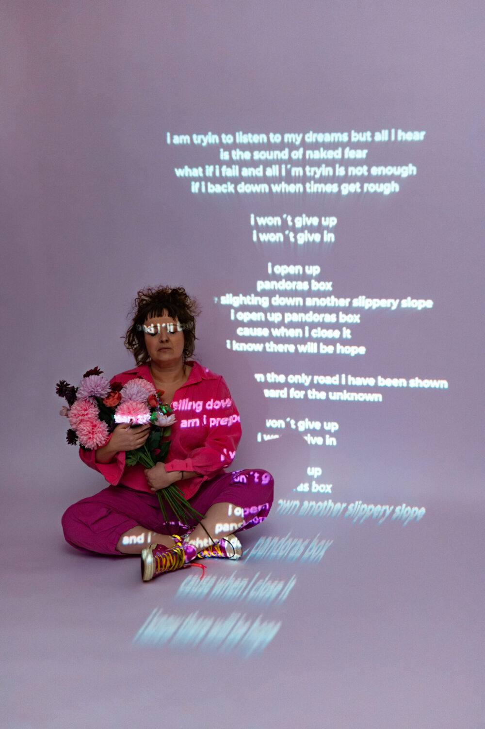 Barbara sitzend mit einem rosa Strauß Blumen in den Händen vor dem projizierten Text von Pandora's Box